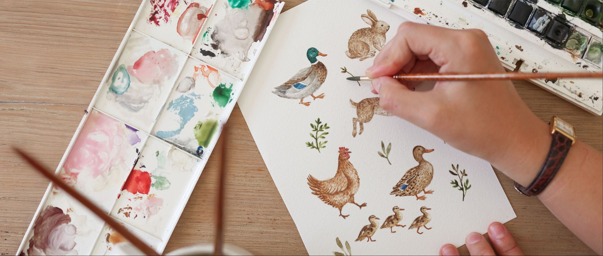 A close-up of a hand holding a paintbrush and painting a watercolor illustration of a duck on a sheet of paper, with a watercolor palette and other art supplies on a wooden table.